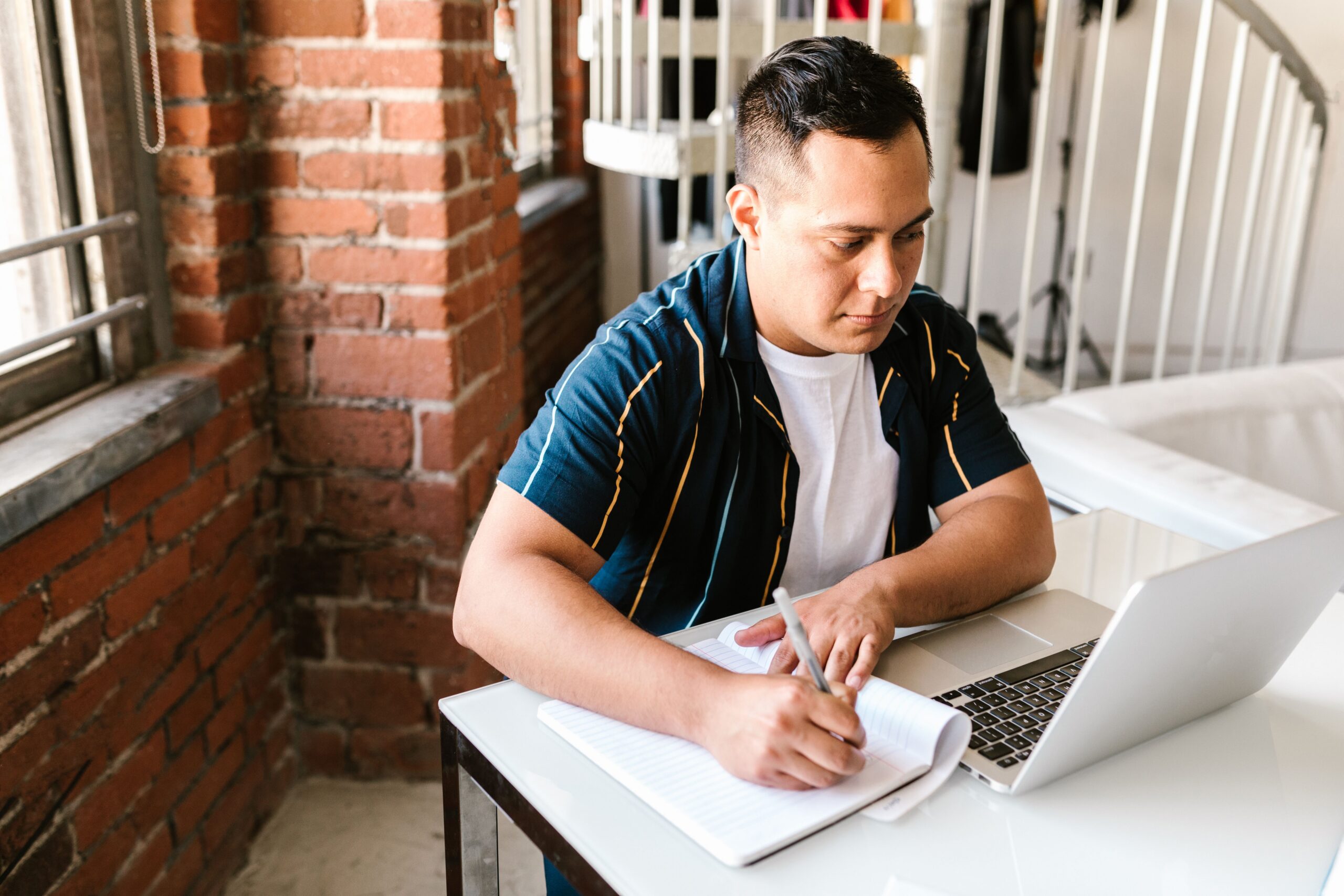 A person making notes in a notebook beside an open laptop