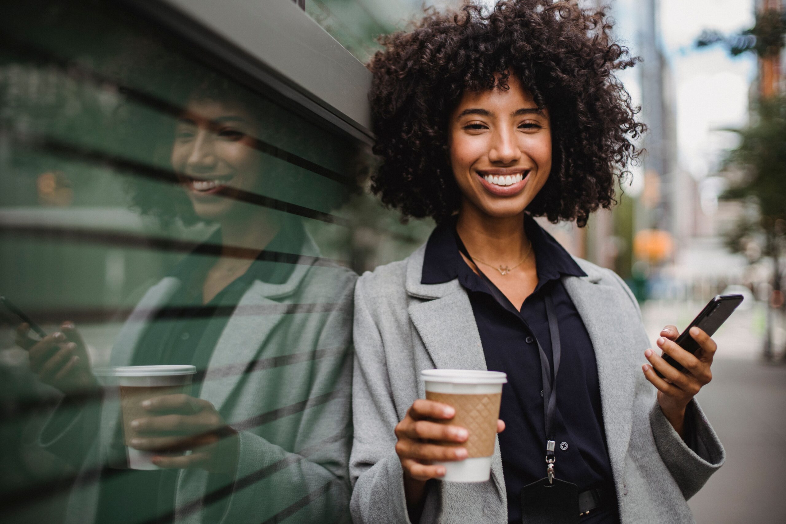 A person smiling outdoors on a city street, holding a coffee and a phone
